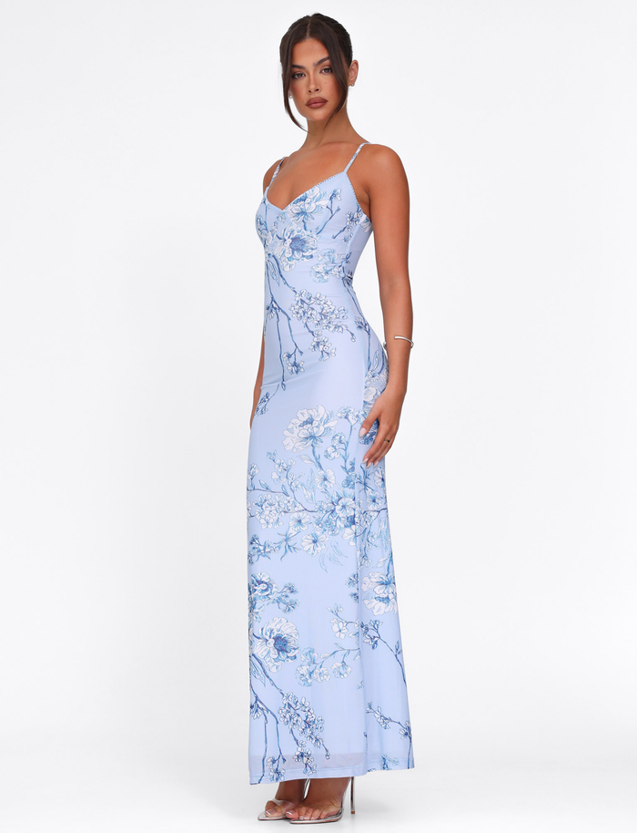 A woman wears the TIGERMIST JULIANNA DRESS in Blue Ceramic Floral—a fitted maxi with spaghetti straps and a V-neckline—paired with clear heels and minimal jewelry, standing against a plain white background.