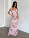 A woman stands indoors against a light wall, wearing the TIGERMIST JULIANNA DRESS - MULTI: COLLAGE: CRANES, a long maxi dress with intricate crane and collage prints. She has long dark hair, strappy heels, and poses elegantly with one hand on her hip.
