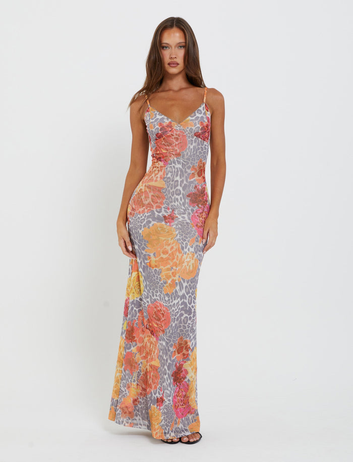 A woman stands against a plain white background wearing the form-fitting, sleeveless JULIANNA DRESS - MULTI : LEOPARD : ORANGE LEOPARD by TIGERMIST. The dress features a vibrant floral and animal print in orange, yellow, and gray tones. She has long, straight hair and is looking directly at the camera.