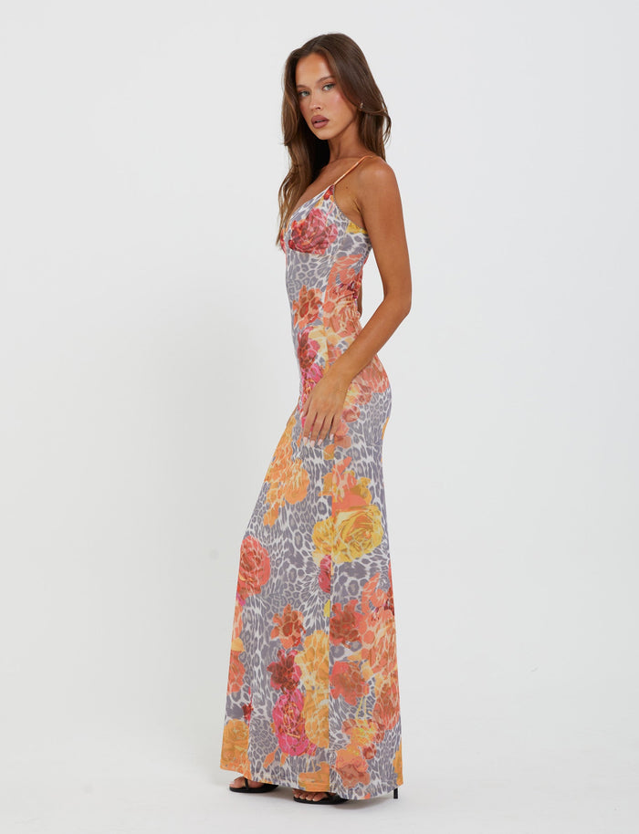 A woman is standing and modeling the Julianna Dress - Multi : Leopard : Orange Leopard by TigerMist. The sleeveless, body-hugging, floor-length dress showcases a vibrant floral and geometric pattern in shades of orange, yellow, pink, and gray. She has long brown hair and is posing sideways with one hand resting on her hip.