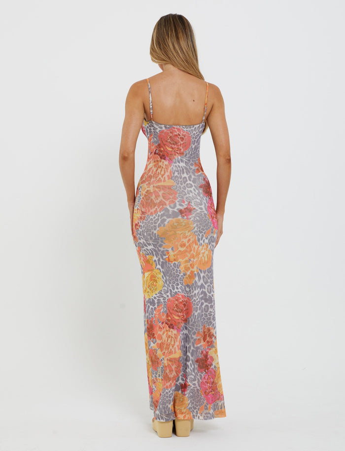 A woman with long hair, facing away from the camera, is wearing the Julianna Dress by TigerMist. The colorful sleeveless maxi dress features an abstract mix of floral and orange leopard print designs. She stands in a relaxed pose against a plain white background.