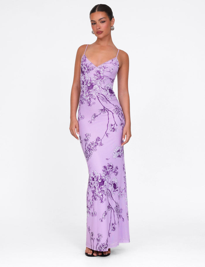 A woman confidently wears the TIGERMIST JULIANNA DRESS in purple ceramic floral, a maxi gown with thin straps, paired elegantly with tied-back hair, hoop earrings, and black heels against a plain white backdrop.