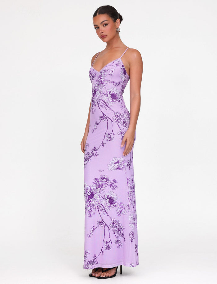 A person in a TIGERMIST JULIANNA DRESS - PURPLE : FLORAL : PURPLE CERAMIC FLORAL, maxi-length lilac design, stands against a plain background. Their hair is styled in an updo with elegant earrings and high heels.