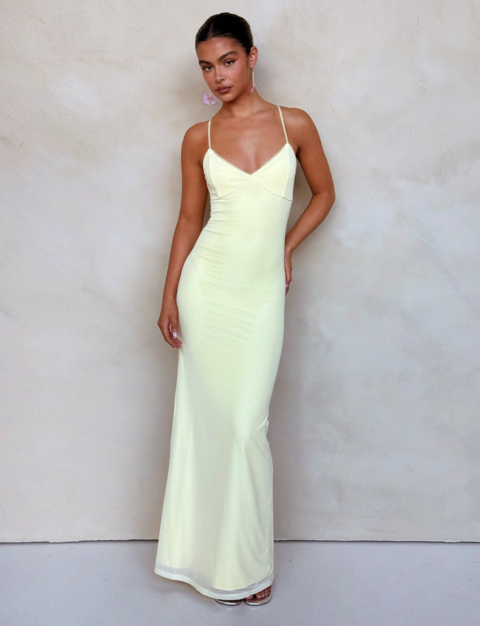 A woman stands against a light-colored background, wearing the form-fitting, floor-length JULIANNA DRESS in LEMON from TIGERMIST, featuring thin spaghetti straps. She has one hand resting on her hip and the other hanging by her side. Her hair is styled back, and she is wearing minimal accessories.