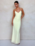 A woman stands against a light-colored background, wearing the form-fitting, floor-length JULIANNA DRESS in LEMON from TIGERMIST, featuring thin spaghetti straps. She has one hand resting on her hip and the other hanging by her side. Her hair is styled back, and she is wearing minimal accessories.