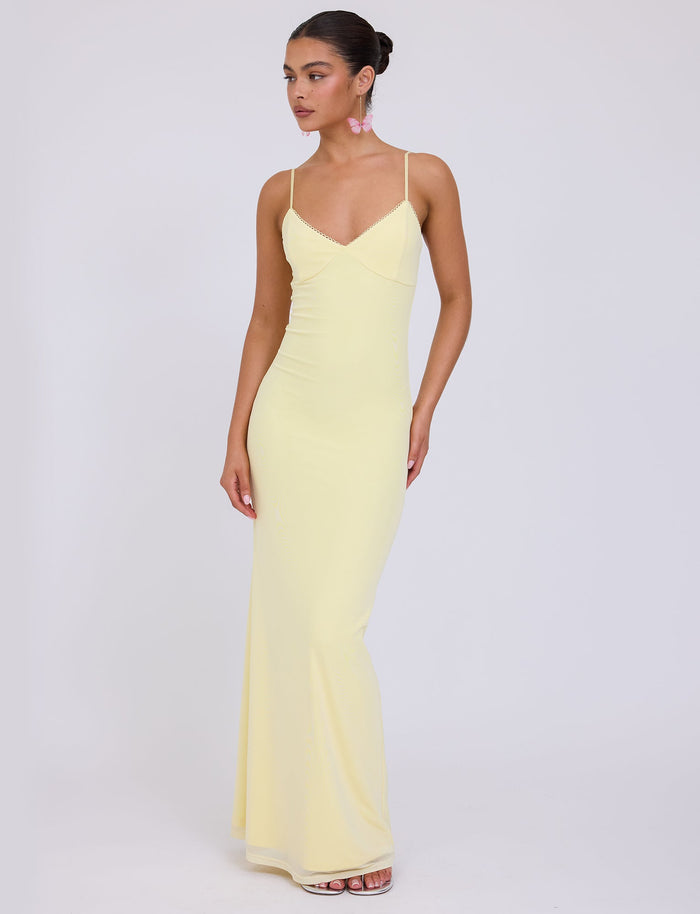 A woman stands against a plain background wearing the form-fitting JULIANNA DRESS - YELLOW : LEMON from TIGERMIST. The dress features thin spaghetti straps and a slight V-neckline. She has her hair tied back and is accessorized with pink earrings, appearing to be looking slightly off-camera.