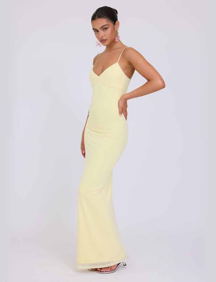 A woman poses in the JULIANNA DRESS - YELLOW : LEMON by TIGERMIST, a floor-length dress with thin shoulder straps and a fitted bodice. She has dark hair pulled back, wears dangling earrings, and stands against a plain white background. She is looking at the camera with one hand on her hip.