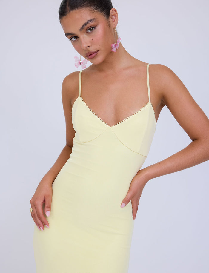 A person with dark hair tied back is wearing the JULIANNA DRESS - YELLOW : LEMON from TIGERMIST and pink butterfly earrings. They are looking slightly downward, with one arm resting on their hip and the other hanging by their side. The background is white.