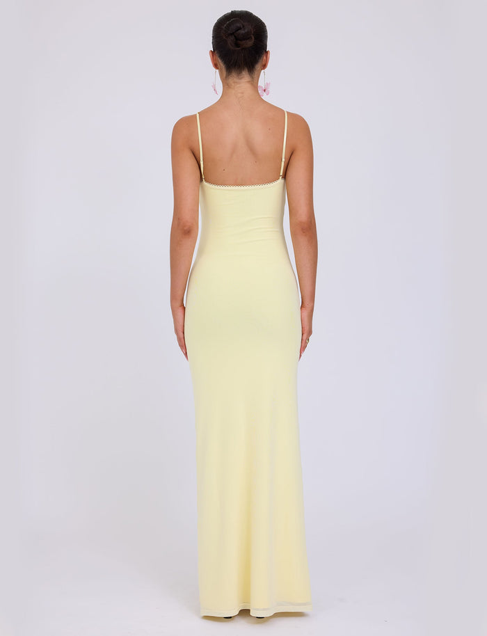 A woman stands with her back to the camera, wearing the JULIANNA DRESS - YELLOW : LEMON by TIGERMIST, a long, fitted dress with thin shoulder straps. Her dark hair is styled in a high bun, and she accessorizes with dangling earrings. The background is plain white.