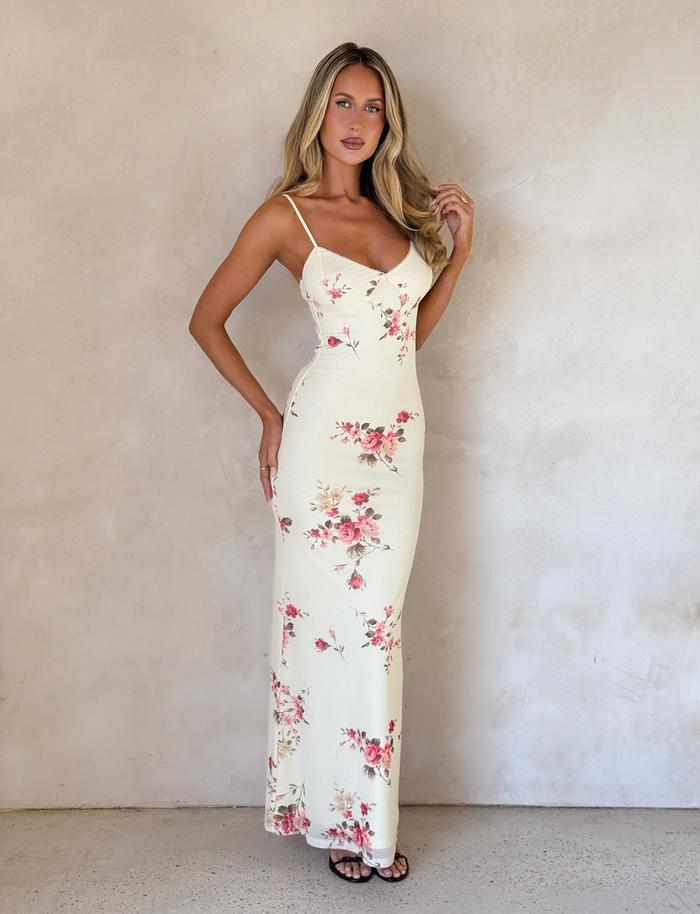 A woman stands against a light wall in the TIGERMIST JULIANNA DRESS in yellow macro pop floral, featuring adjustable spaghetti straps, complementing her long blonde hair and black sandals as she gently touches her hair.