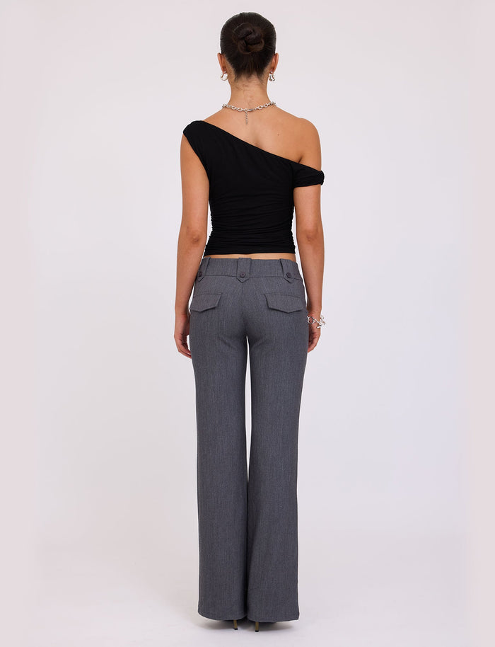 A person stands with their back to the camera, dressed in the KADENCE TOP - BLACK and high-waisted, grey wide-leg trousers from TIGERMIST. Their hair is styled in an updo and adorned with a necklace and bracelets. The background features a plain, light-colored backdrop.