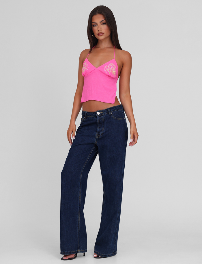 A woman stands confidently against a plain background, wearing the TIGERMIST KAELAH TOP in hot pink, paired with high-waisted wide-leg dark blue jeans and black open-toe heels.