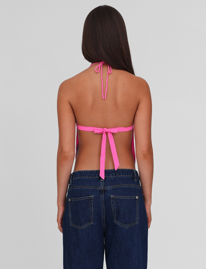 A woman with long brown hair is seen from the back wearing the TIGERMIST KAELAH TOP in hot pink, an open-back halter style tied at the neck and back, paired with dark blue jeans. The background is plain light gray.