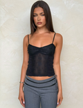 A woman with straight brown hair wears the TIGERMIST KALLIE TOP in black, featuring adjustable straps, paired with gray pants. She stands against a light textured wall, looking at the camera with a neutral expression.