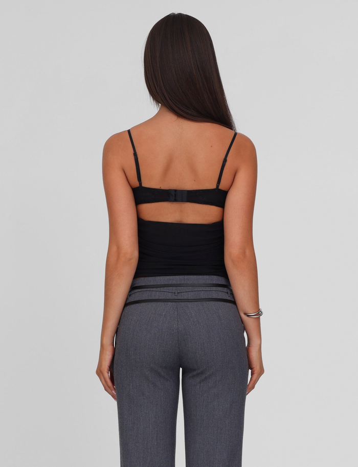 A woman with long straight brown hair, wearing the TIGERMIST KALLIE TOP - BLACK with adjustable straps and gray pants, stands facing away from the camera against a plain light background.