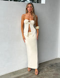 A woman stands against a blank wall, wearing a strapless white top with a front tie and TIGERMIST's KAROLINA JERSEY SKIRT in ivory. She has long, straight hair and accessorizes with wooden bangles on one wrist, complemented by brown sandals. She gazes confidently at the camera.