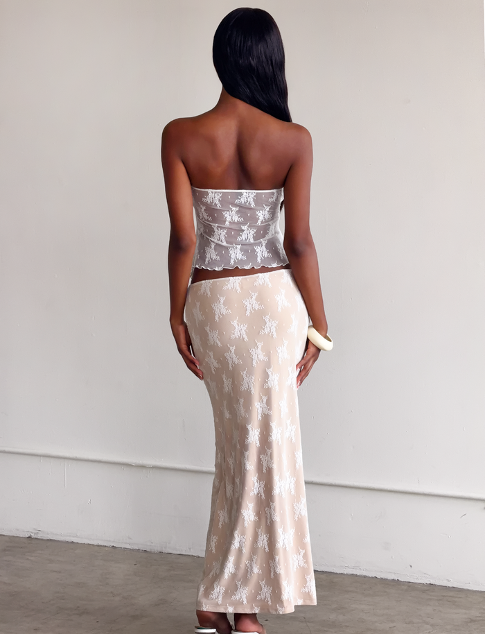 A woman with long straight hair stands indoors facing away, wearing the TIGERMIST KAROLINA TOP in white lace and holding a small clutch, set against a plain light-colored wall.