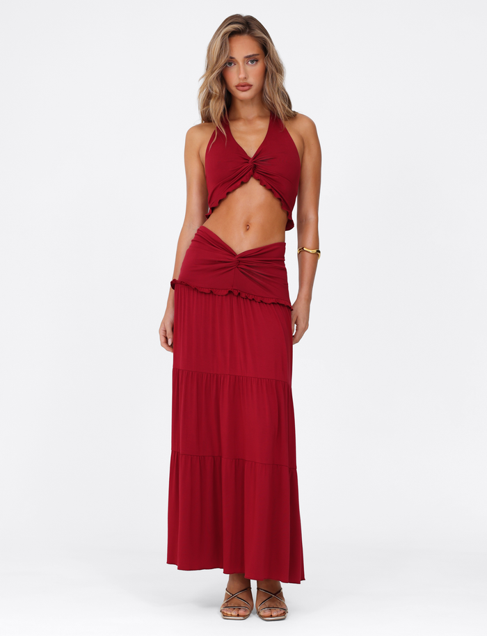 A woman wears the TIGERMIST KATYA HALTER TOP in deep red, featuring a crop length and front twist detail, paired with a matching tiered maxi skirt. She stands facing forward with loose wavy hair, gold sandals, and a neutral expression.