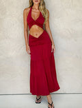 Against a textured wall, a person wears TIGERMIST's KATYA MAXI SKIRT in red with tiered panels and a sleeveless halter top featuring a front twist. The wide waistband enhances their silhouette, complemented by black sandals.