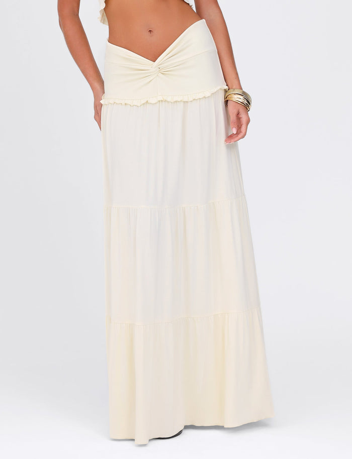 A person wearing the KATYA MAXI SKIRT - WHITE : CREAM by TIGERMIST, featuring a knotted waistband and tiered design for comfort with its high stretch fabric, accessorized with multiple gold bangles against a plain white background.