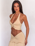 A woman with long dark hair wears the TIGERMIST KATYA TOP in tan, featuring a halter neck and twist detail. She stands against a plain light background, looking confidently at the camera.
