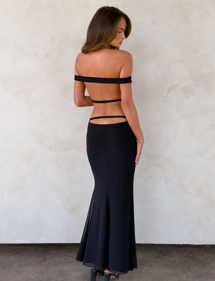 A woman faces away, wearing the KERRIGAN MAXI DRESS - BLACK by TIGERMIST, featuring an off-shoulder design, open back, and thin straps accentuating her fitted bodice and waist against a light textured wall.
