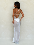 A woman faces away in the TIGERMIST KIAH MAXI DRESS - WHITE : ICE, featuring a sheer style with criss-cross shoulder straps and an open back, set against a light textured wall.