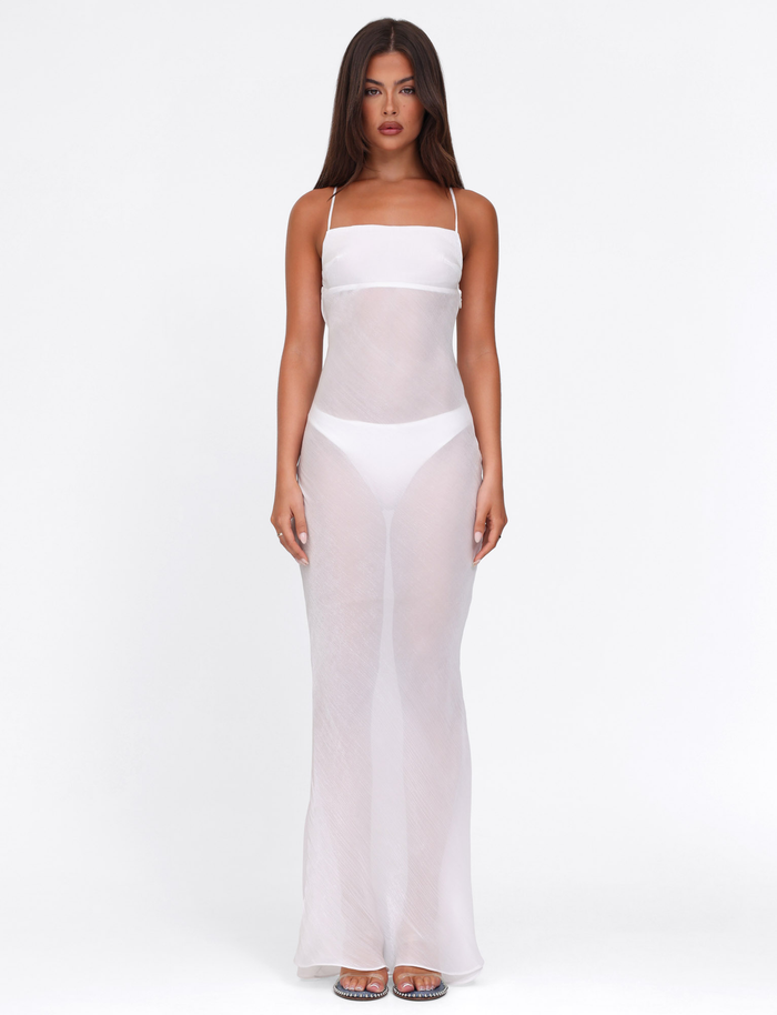 A woman faces forward in the TIGERMIST KIAH MAXI DRESS - WHITE : ICE, featuring sheer fabric and crisscrossed shoulder straps over white undergarments and clear heels, set against a plain white background.