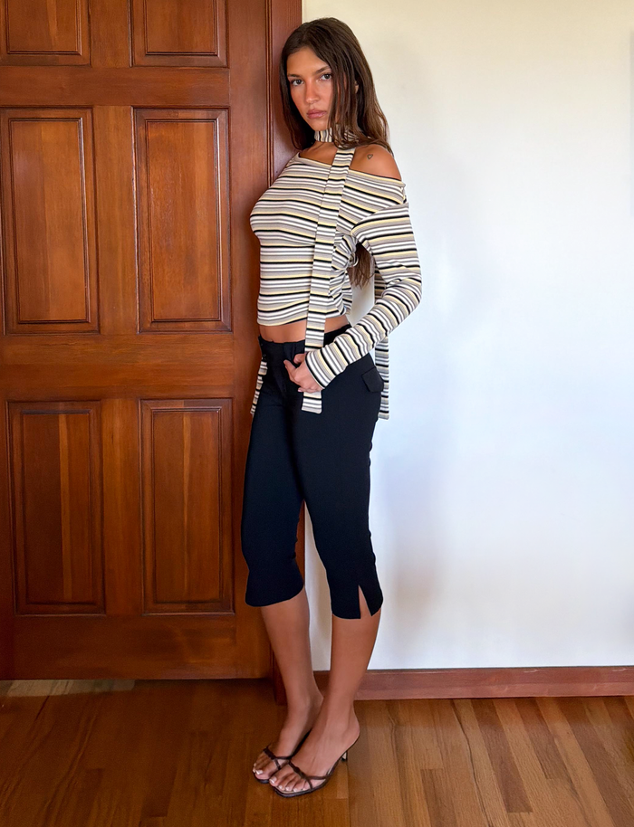 A woman stands indoors by a wooden door, wearing the TIGERMIST KIERSTEN SCARF TOP - MULTISTRIPE, paired with black cropped pants and heeled sandals. She poses with one hand in her pocket, looking at the camera neutrally.