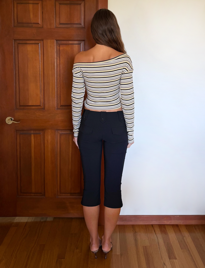 A woman with long, wavy brown hair wears the TIGERMIST KIERSTEN SCARF TOP - MULTISTRIPE, paired with black cropped pants and transparent heels. She stands indoors on a hardwood floor by a wooden door and a white wall.