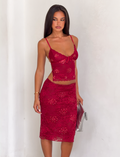 A woman with long, straight dark hair stands against a white wall, wearing the TIGERMIST KIRRA BEADED SEQUIN LACE MIDI SKIRT in red and a sleeveless red lace crop top. She holds a silver clutch.