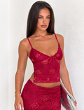 A woman with long dark hair wears the TIGERMIST KIRRA BEADED SEQUIN LACE TOP in red with a matching skirt, holding a silver clutch bag, standing against a plain white wall.