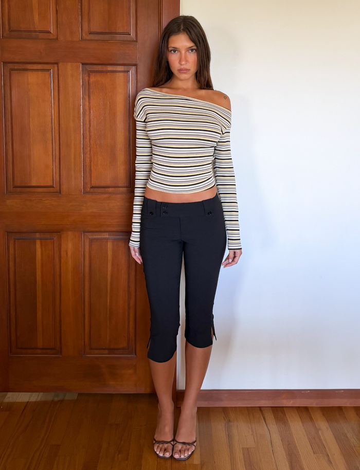 A woman stands indoors before a wooden door, wearing TIGERMIST's striped off-the-shoulder long-sleeve top, KITTIE CAPRI - BLACK pants, and clear-strap heels. She faces the camera with a neutral expression.