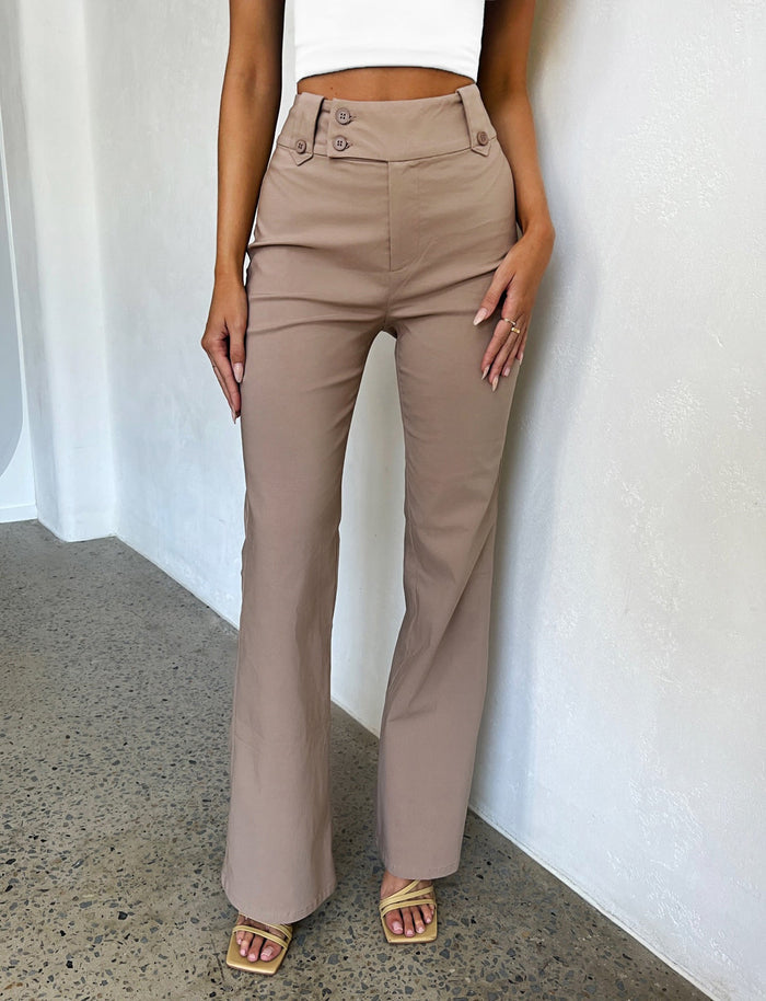 A person dressed in the KITTIE HIGH RISE PANT - BEIGE : TAN from TIGERMIST, showcasing button details at the waistline. The individual is complemented by a TIGERMIST white crop top and matching beige open-toe sandals, standing on a polished concrete floor against a pristine white wall.
