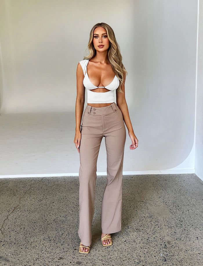 A woman with long blonde hair stands facing the camera against a minimalist indoor background. She is wearing a white cropped top from Tiger Mist and the Kittie High Rise Pant in beige from TIGERMIST, which features high-waisted design and flared legs. She has on open-toed heels, her left hand rests at her side while her right arm is slightly bent.