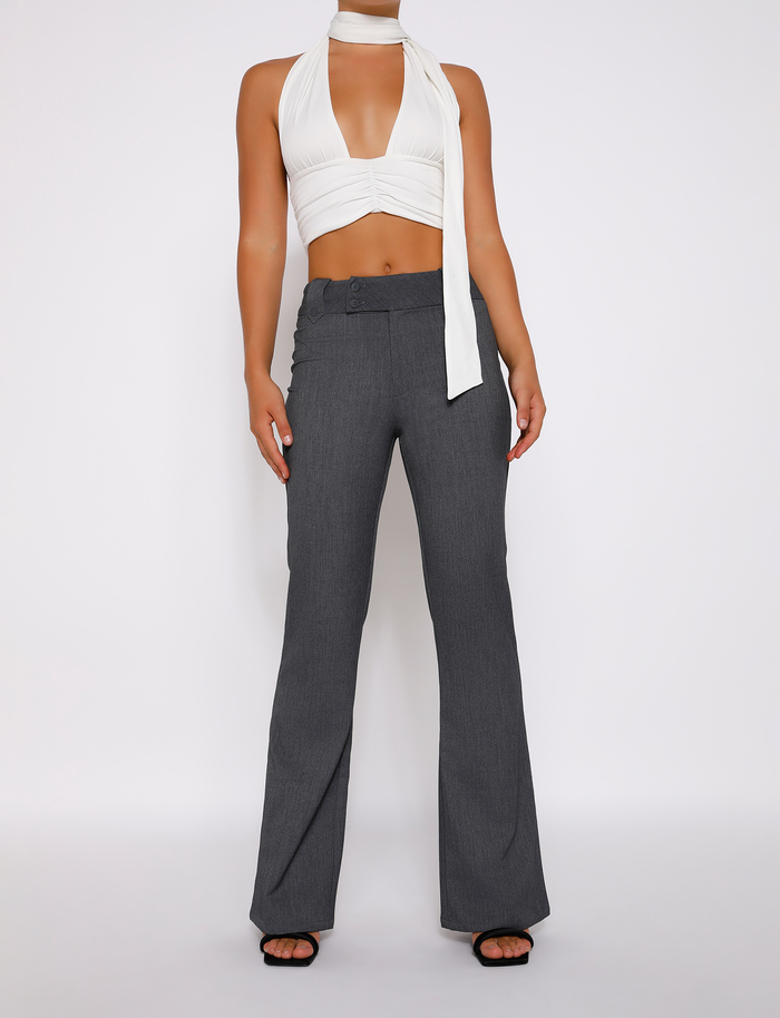 A person stands against a white background wearing a sleeveless white halter crop top, TIGERMIST's KITTIE HIGH RISE PANT - GREY with a flared leg and asymmetrical waistband, and black open-toe heels.