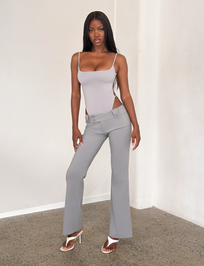 A woman poses confidently indoors against a white wall, wearing a fitted light gray tank top and the TIGERMIST KITTIE PANT - FROST GREY, featuring hip cutouts. She completes the look with white heeled sandals and loose dark hair.