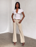 A woman stands indoors in TIGERMIST’s KITTIE PANT - SAND, crafted from stretch suiting fabric, paired with a fitted white short-sleeve top and black heeled sandals against a light wall and speckled floor.