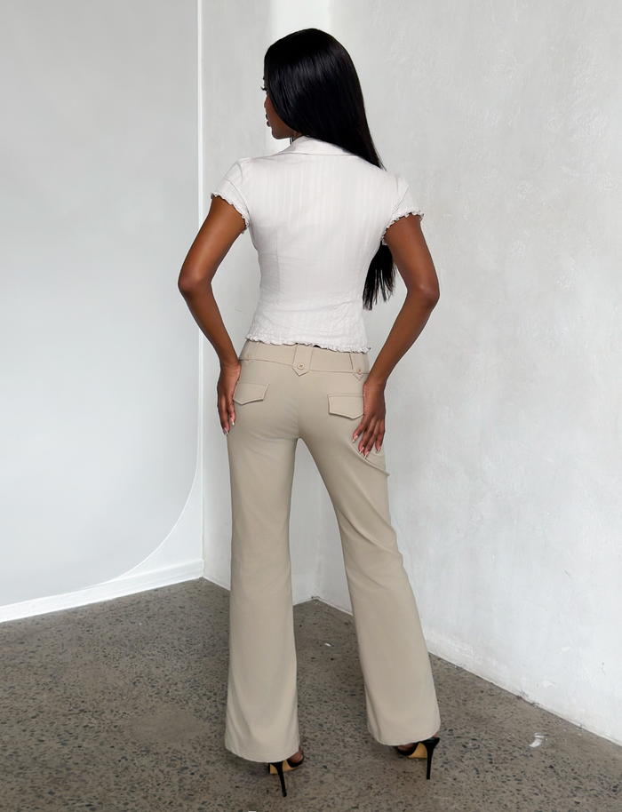 A woman with long dark hair stands facing a white textured wall, wearing a short-sleeve white blouse, TIGERMIST KITTIE PANT in sand stretch suiting fabric, and black high heels. The photo is taken from behind on a speckled floor.