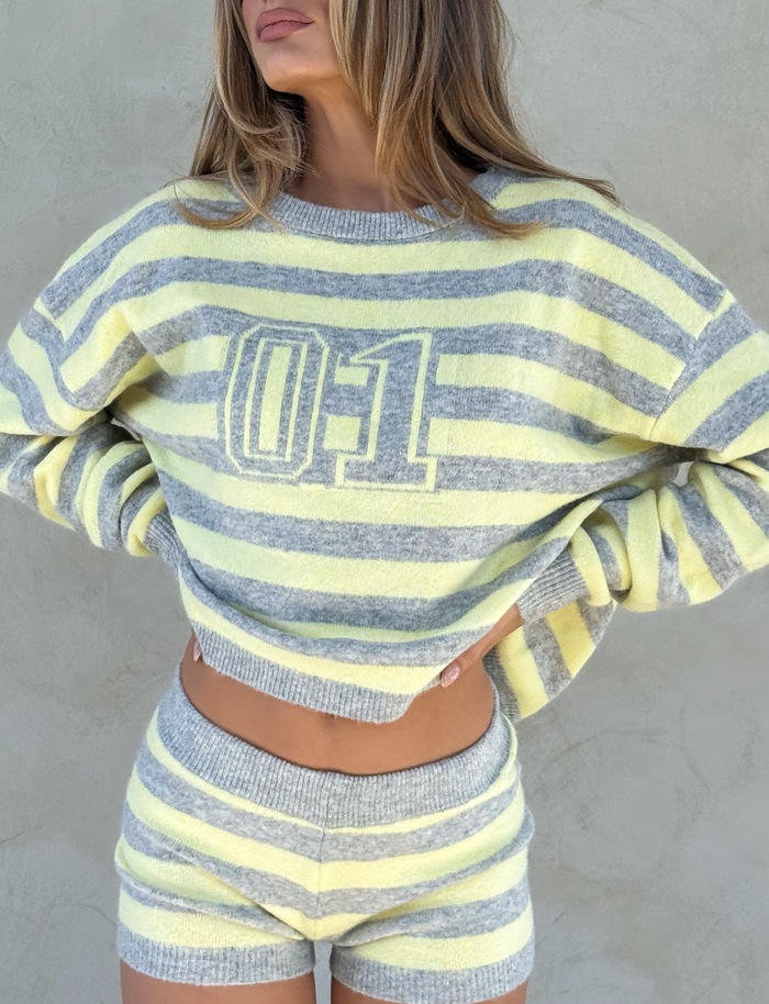 A woman stands against a neutral wall wearing the TIGERMIST KOA JUMPER in yellow and grey stripes, featuring an oversized fit with a sporty "01" print on the front, paired with matching striped knit shorts.