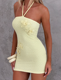 A woman wears the TIGERMIST KORRI DRESS - BUTTER, a strapless light yellow knit mini dress with crochet floral appliques and halter straps, standing against a textured gray wall.