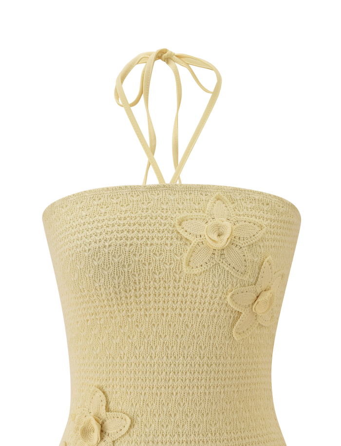 The KORRI DRESS - BUTTER by TIGERMIST features delicate crochet details and a soft beige hue, with thin neck ties and floral appliques adding texture and charm to the front.