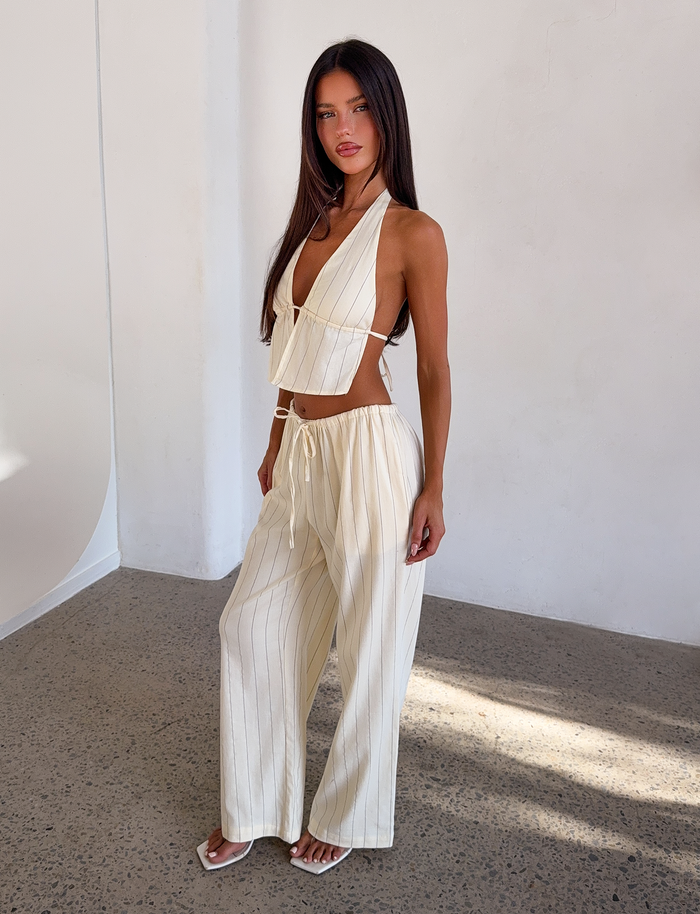 A woman poses indoors against a white wall wearing the TIGERMIST LAINEY PANT in beige stripe, styled with a cream backless halter crop top, white sandals, and sporting long dark hair.