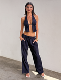 A woman stands against a plain light wall wearing Tiger Mist USA’s CL - LAINEY PANT, a dark blue pinstriped wide-leg style, paired with a matching halter top, white sandals, and a gold watch. She has her hands in her pockets.