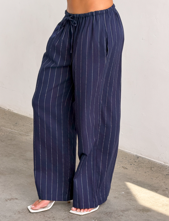 Someone wearing the TIGERMIST LAINEY PANT in navy pinstripe—loose-fitting linen pants with an elastic waist—paired with white sandals, stands on a concrete floor against a light-colored wall.