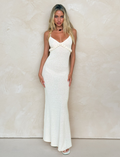 A woman with long blonde hair wears the TIGERMIST LAVELLE MAXI DRESS in white/cream with adjustable criss cross straps and white sandals, standing against a light beige wall and looking directly at the camera.