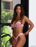 A woman with long, wavy hair stands by a large window wearing the LAVINA SET - RED : SALSA from TIGERMIST. One hand rests on her hip while the other is placed against the window frame. The background shows lush green foliage outside.