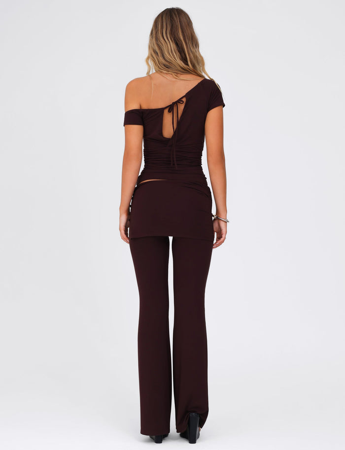 A woman with long, wavy hair is wearing the TIGERMIST dark-brown, off-the-shoulder top featuring a back tie detail and the LEILANI PANT - BROWN : CHOCOLATE flared leg pants made of high stretch fabric. She stands on a plain white background, facing away from the camera.