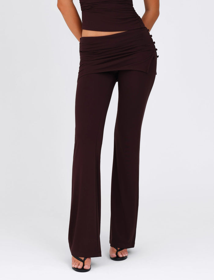 A person dressed in the LEILANI PANT - BROWN: CHOCOLATE from TIGERMIST, crafted from high-stretch fabric and featuring a flared design, paired with a matching asymmetric wrap skirt. They complete the look with open-toed black sandals against a plain white background.