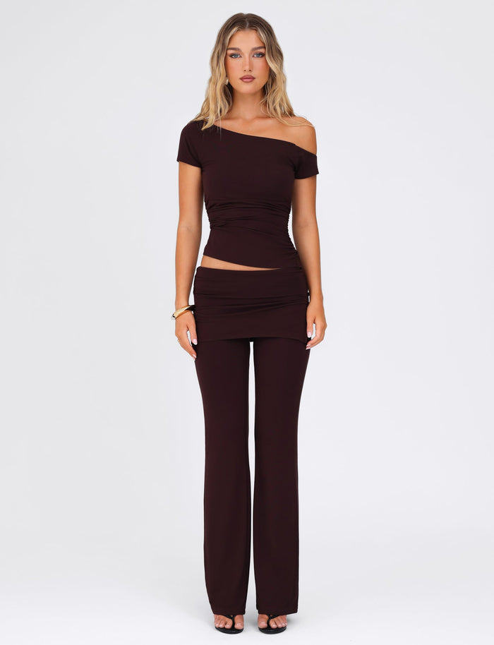 A stylish individual poses against a simple background wearing the LEILANI PANT – BROWN : CHOCOLATE by TIGERMIST, featuring a flared leg design. They pair it with an elegant, off-shoulder black top that highlights the high stretch fabric and reveals a glimpse of their midriff. With long, wavy hair cascading over their shoulders, they complete the look with heels.